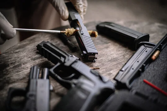 Gloved hands cleaning a pistol slide with a brass brush, with pistol frames and magazines on the bench
