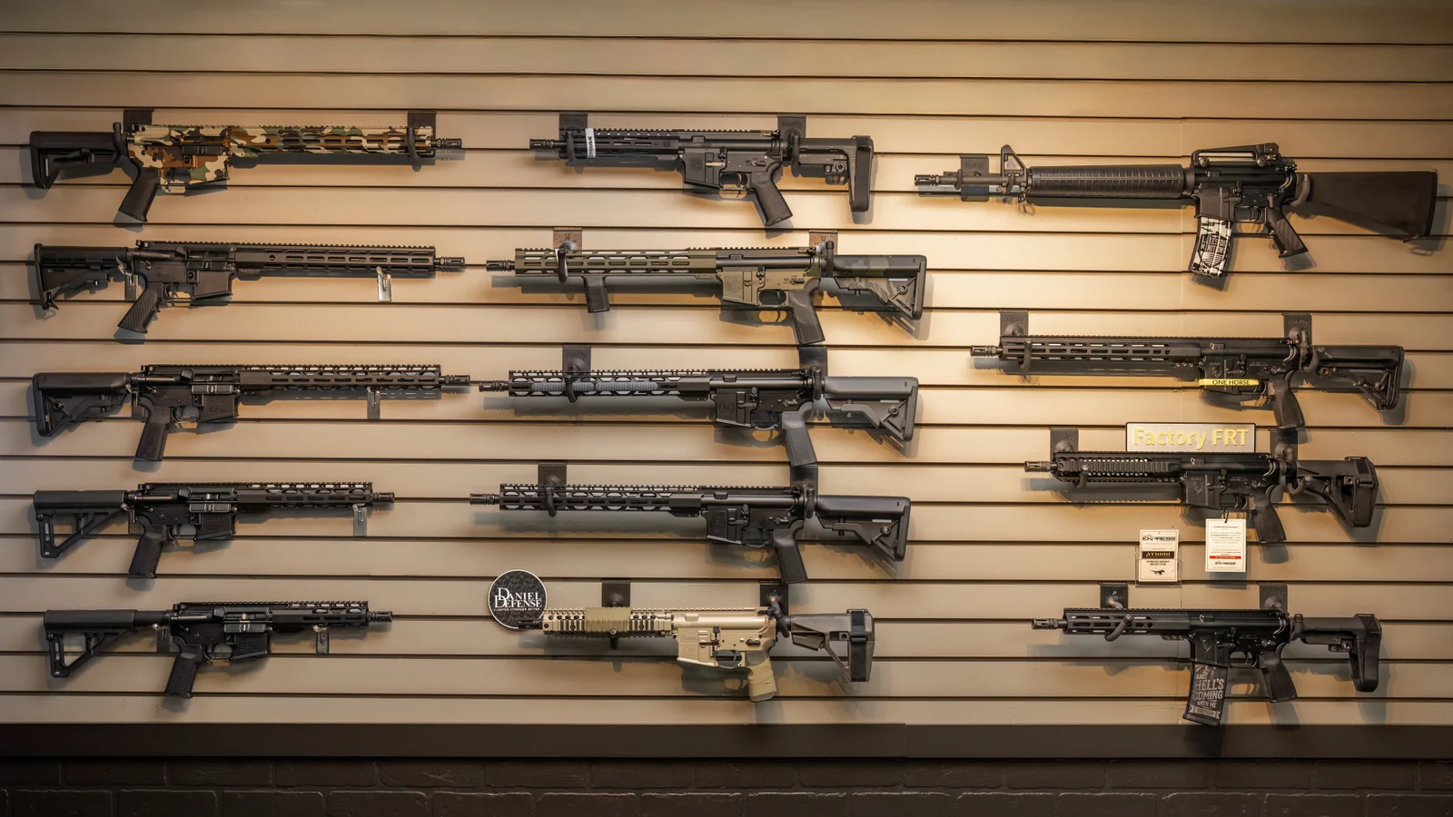 Slatwall of AR-style rifles on hooks in rows, with tan, black and camo finishes