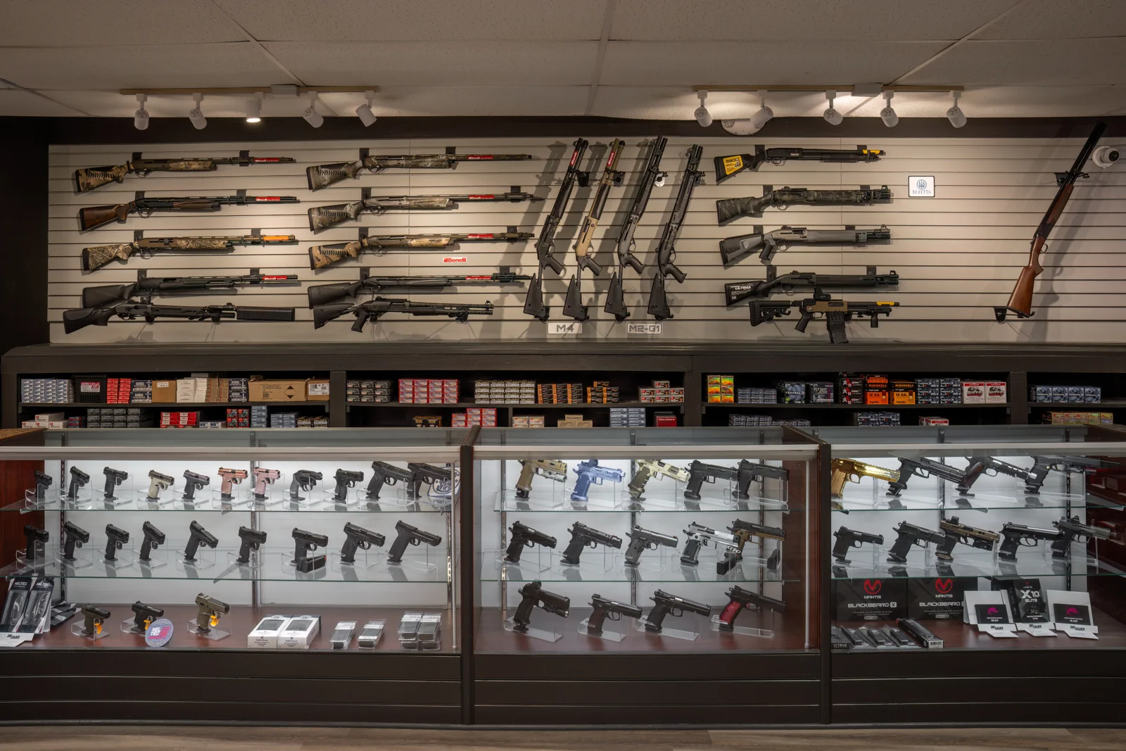 Shotgun and rifle wall above glass cases of handguns, with boxes on the lower shelf