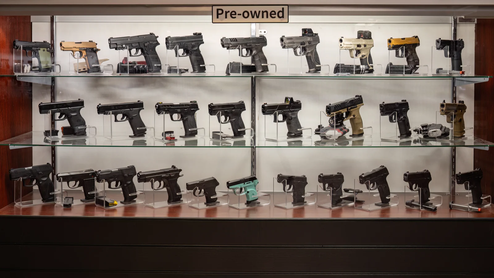 Glass case of pre-owned handguns on three shelves under a Pre-owned sign