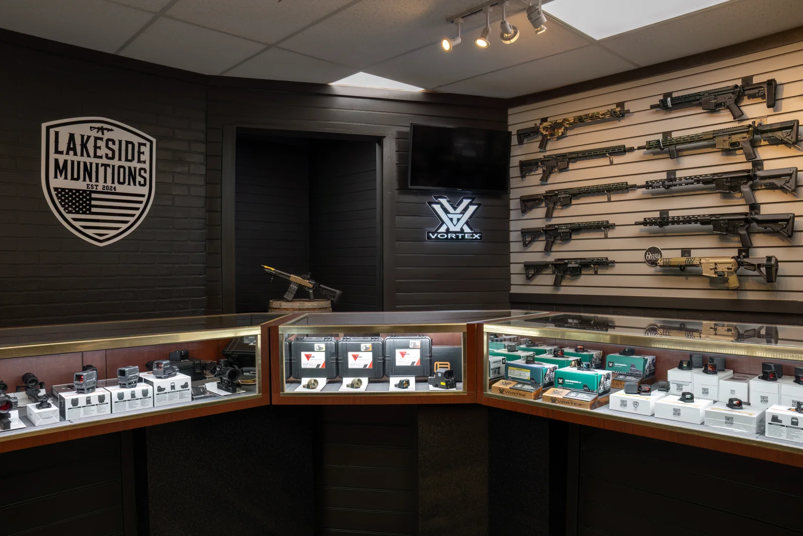 Corner with a Lakeside shield logo, a lit Vortex sign, optics cases and an AR-style rifle wall