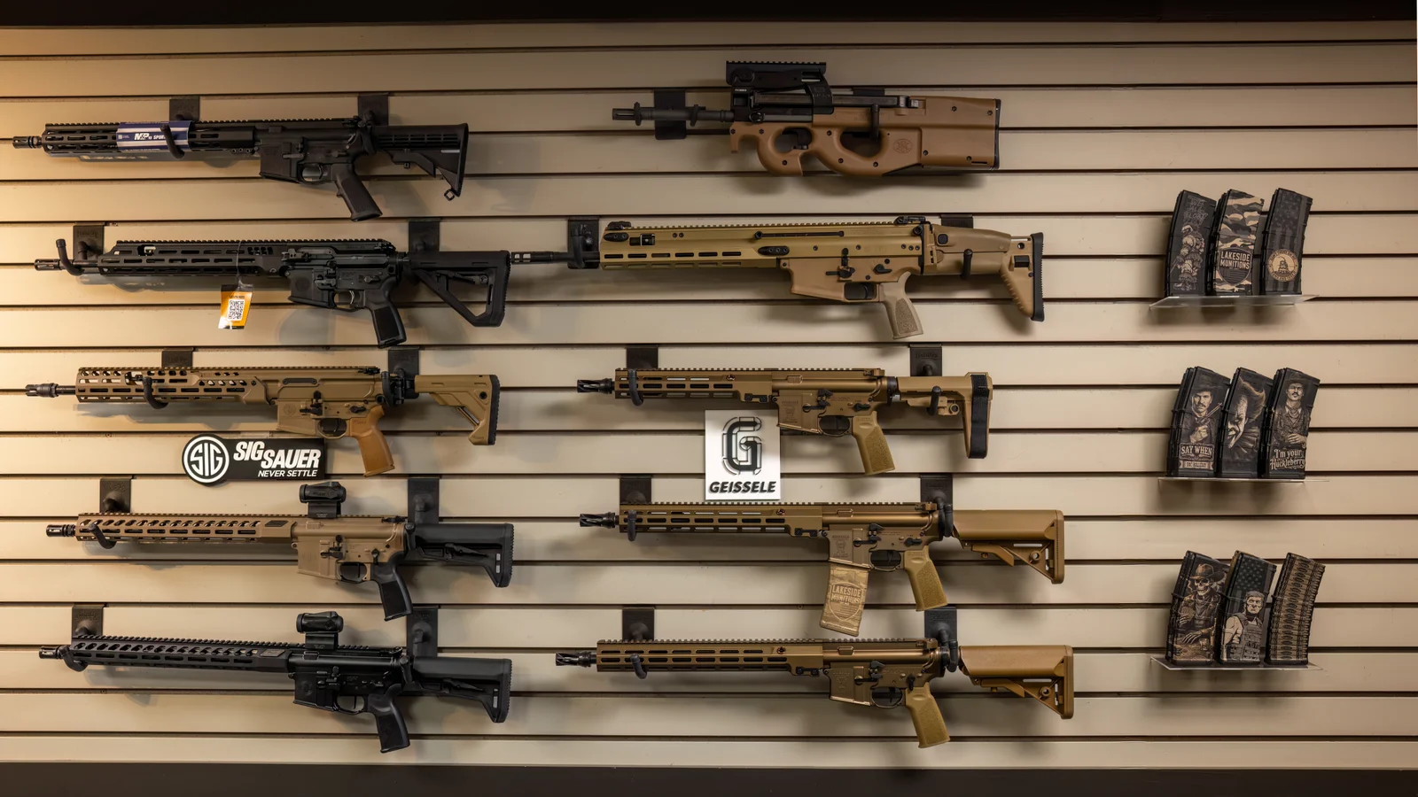 Slatwall of tan and black AR-style rifles with Sig Sauer and Geissele signs, and magazines on the right
