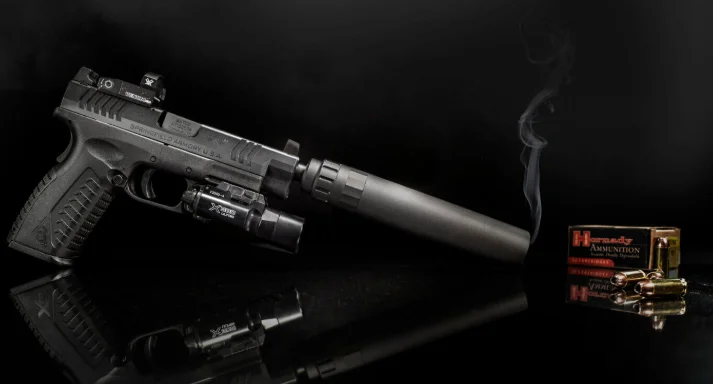 Black pistol with a red dot, weapon light and suppressor beside a box of Hornady ammunition